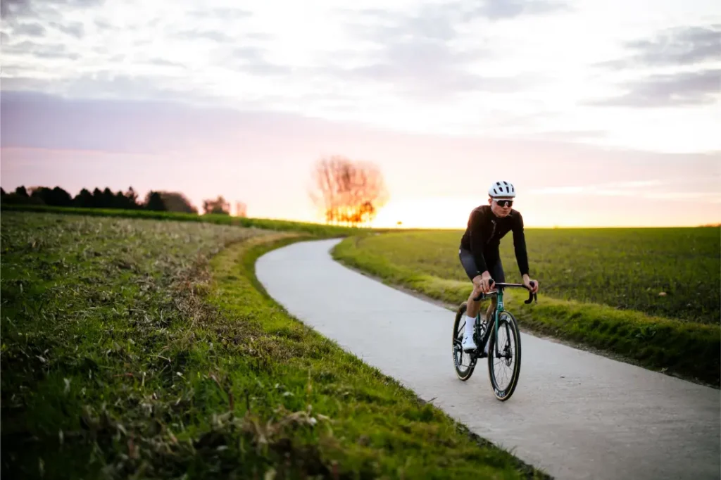 Un ciclista con una Casco Lazer Sphere Kineticore recorre una sinuosa carretera rural al atardecer, rodeado de campos verdes y un cielo despejado.