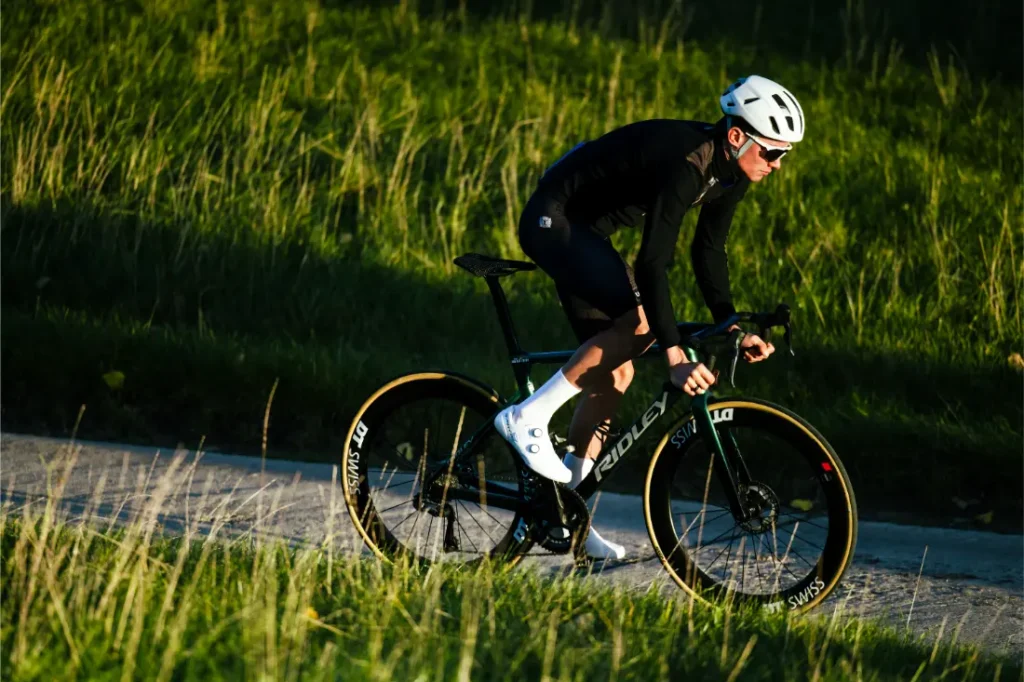 Un ciclista con equipación negra y casco Casco Lazer Sphere Kineticore monta en bicicleta de carretera por un camino pavimentado a través de un terreno cubierto de hierba a la luz del día.