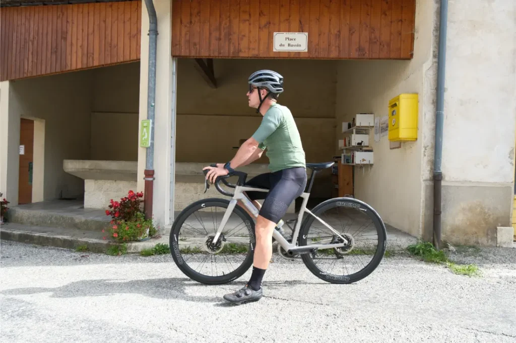 Un ciclista con casco y maillot verde permanece de pie junto a una moderna bicicleta de carretera equipada con Corima Essentia 42 ruedas endurance frente a un edificio rústico con un cartel en el que se lee "Place du Bascil".