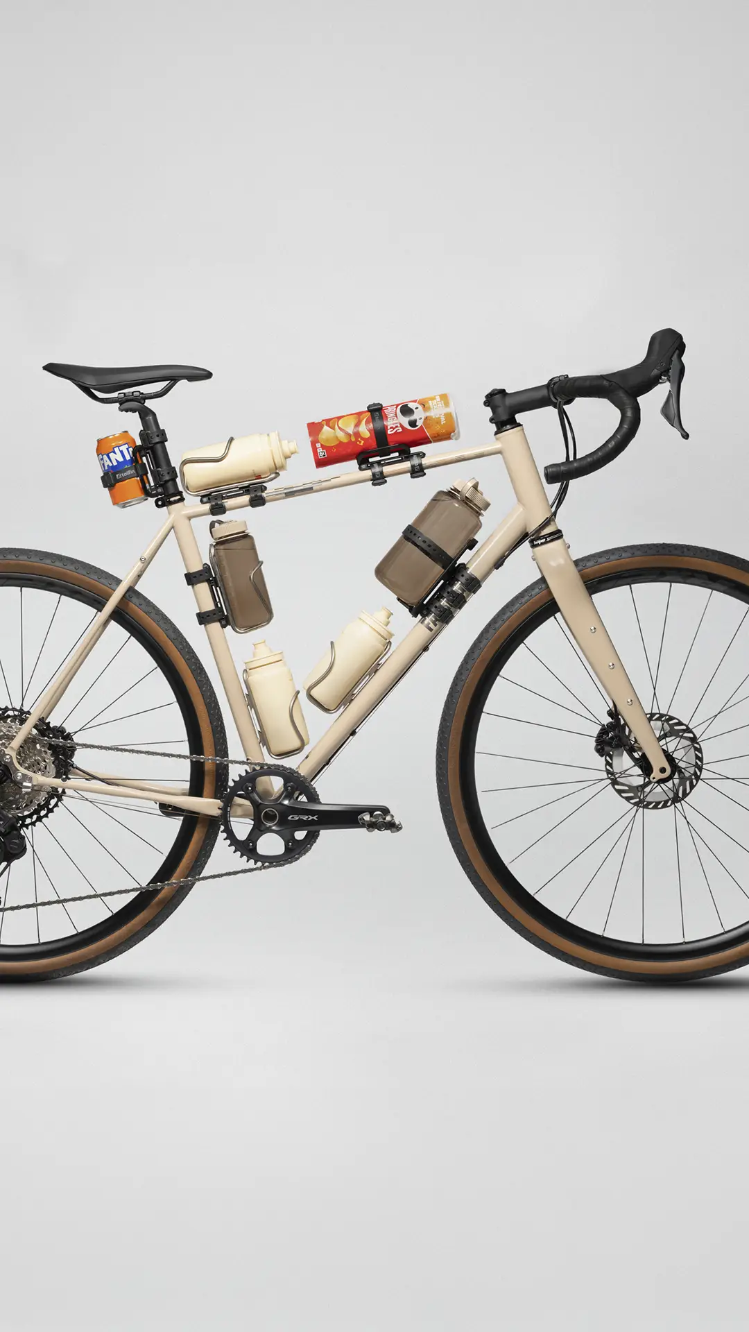 Una bicicleta de grava beige con múltiples portabidones sostiene botellas de agua, una lata de Fanta y otra de Pringles sobre un fondo blanco liso.