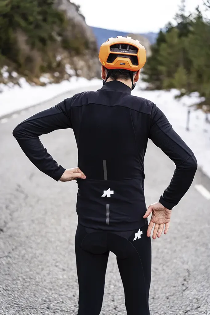 Ciclista con traje negro y casco naranja -con la chaqueta extrema ASSOS- de pie en una carretera de montaña con nieve sobre los hombros, de espaldas a la cámara.
