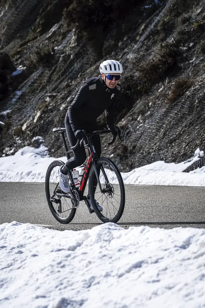 Un ciclista con una chaqueta extrema conduce una bicicleta de carretera cuesta arriba por una carretera asfaltada con nieve a ambos lados y una ladera rocosa y nevada al fondo.