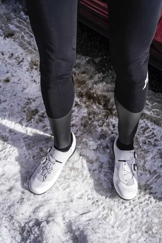 Persona vestida con leggings negros, zapatillas de ciclismo blancas y una Chaqueta ASSOS EQUIPE RS Johdah Ultraz de pie sobre suelo cubierto de nieve junto a un vehículo rojo.