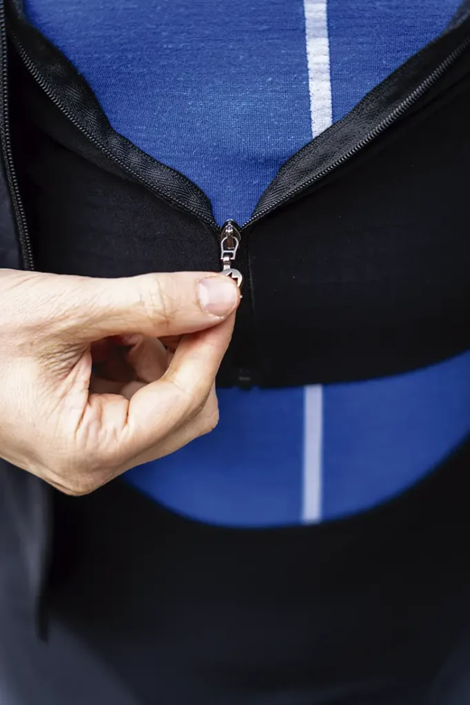 Primer plano de la mano de una persona subiendo la cremallera de una chaqueta negra EQUIPE RS Johdah Ultraz ASSOS sobre una camisa de rayas azules y blancas.