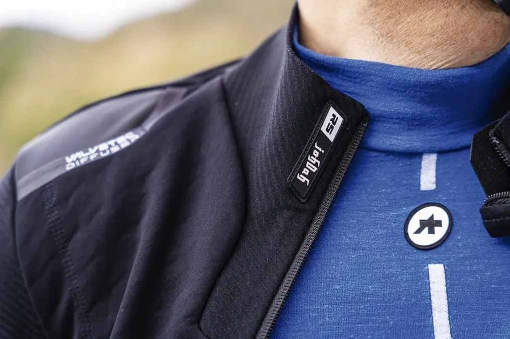 Primer plano de una persona que lleva un maillot de ciclismo azul con el logotipo visible y una chaqueta ASSOS Johdah Ultraz S11 con cremallera parcial, mostrando el texto "SPYKER 2S" en el cuello.