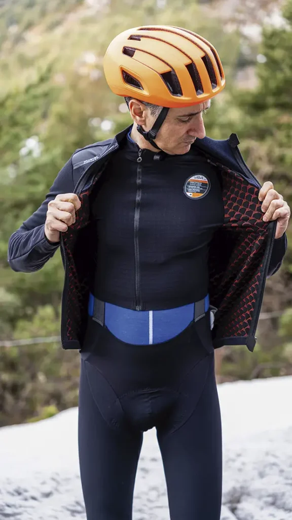 Un hombre con un casco de ciclismo naranja y ropa de ciclismo negra está de pie al aire libre, abriendo la cremallera de su EQUIPE RS Johdah Ultraz chaqueta ASSOS para revelar un forro interior estampado.