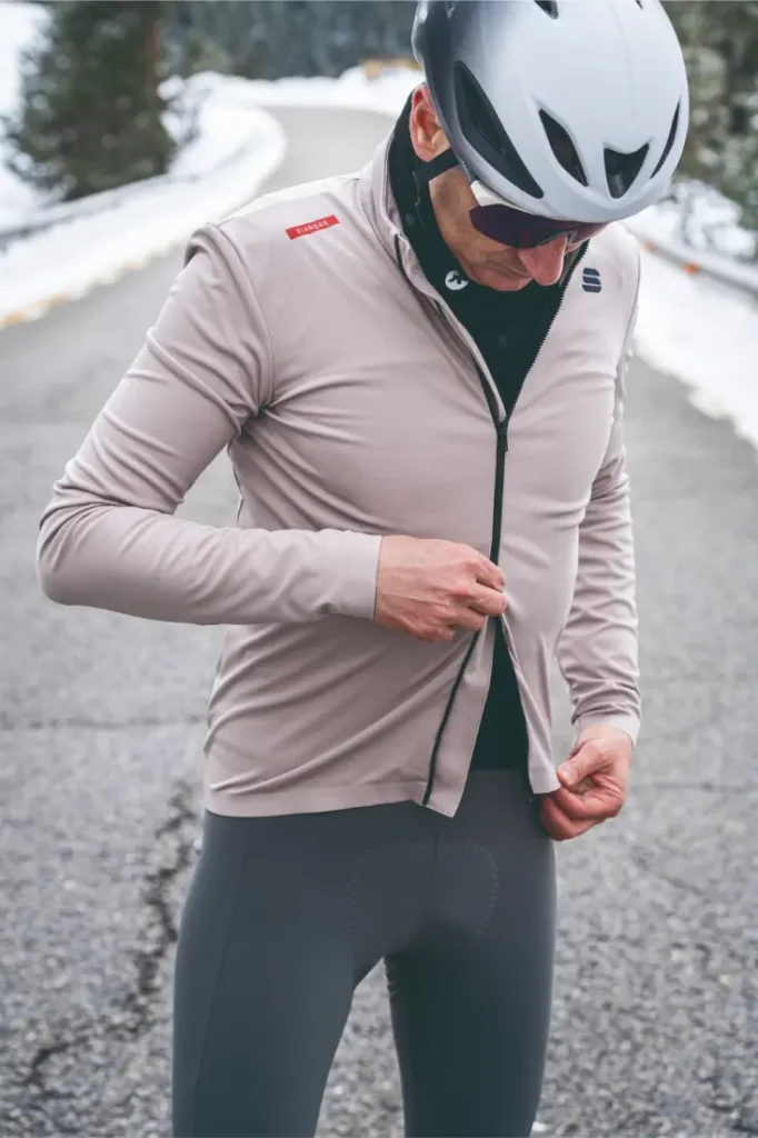 Un ciclista con casco, gafas de sol y la Chaqueta Sportful Fiandre sube la cremallera de su chaqueta en una carretera asfaltada con nieve visible en los laterales, asegurando protección y transpirabilidad en condiciones de frío.
