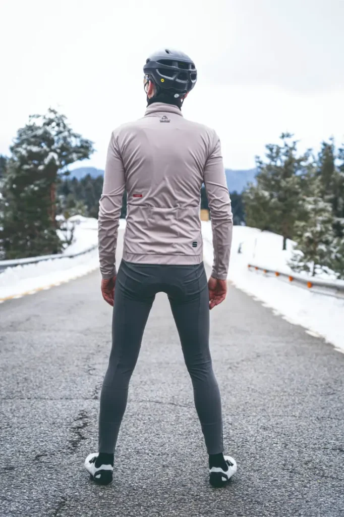 Una persona vestida con casco, camiseta de manga larga y mallas de ciclismo -equipada con una Chaqueta Sportful Fiandre para protección y transpirabilidad- se sitúa en una carretera nevada de espaldas a la cámara.