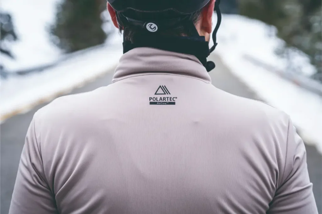 Persona con una Chaqueta Sportful Fiandre de color claro y un casco de ciclista, de pie en una carretera bordeada de nieve con árboles al fondo, lista para la aventura con protección y transpirabilidad en cualquier clima.
