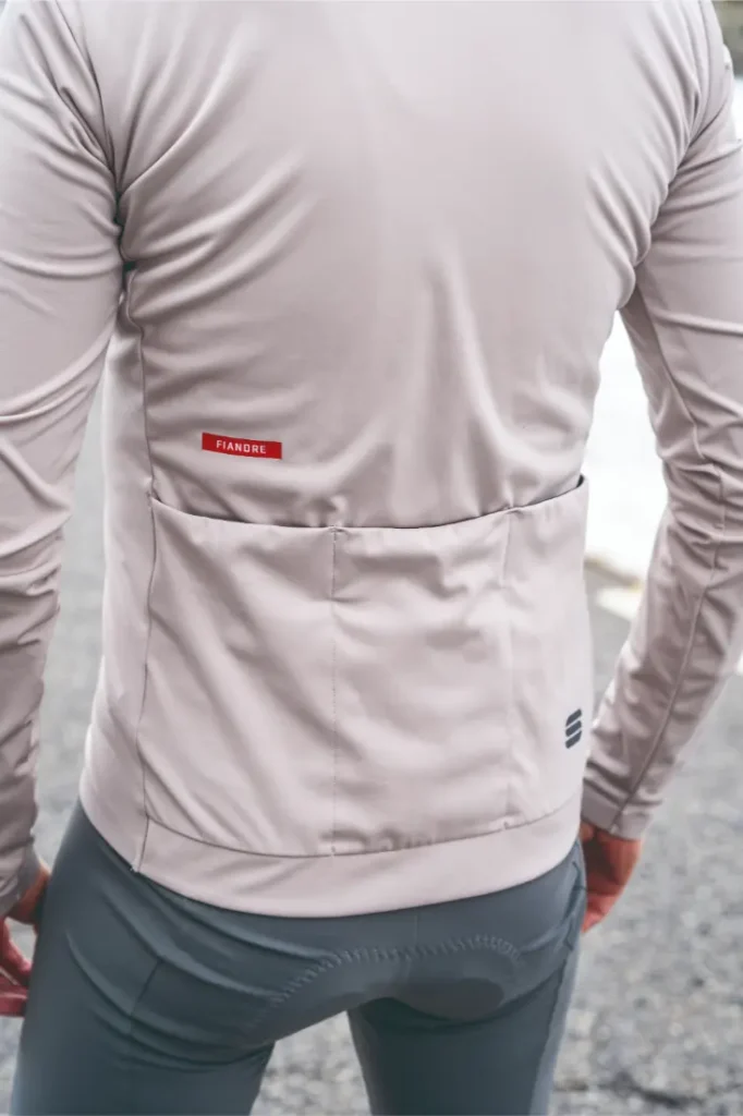 Persona que lleva una Chaqueta Sportful Fiandre de manga larga con bolsillos traseros de color gris claro, disfrutando de la protección y transpirabilidad mientras está de pie al aire libre sobre una superficie pavimentada.