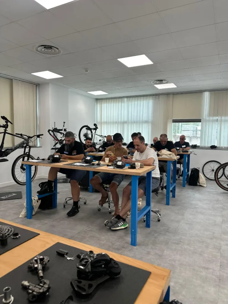 Personas sentadas en bancos de trabajo montando o reparando piezas de bicicleta en una sala luminosa con bicicletas, equipamiento ciclista y materiales de Formaciones Shimano 2026 a la vista.
