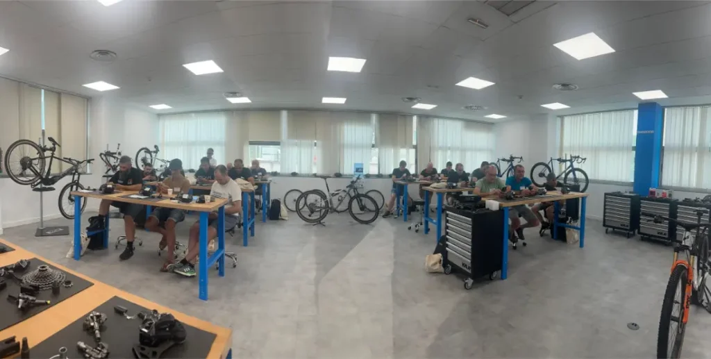 Durante una de las sesiones de Formaciones Shimano 2026, varias personas están sentadas en bancos de trabajo en un luminoso taller, montando o reparando bicicletas con diversas piezas y herramientas visibles por toda la sala.