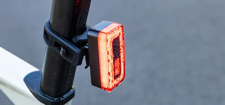 Un piloto trasero rojo Sigma Nyte 70 se sujeta a la tija del sillín de una bicicleta con una correa negra, sobre un fondo gris liso.