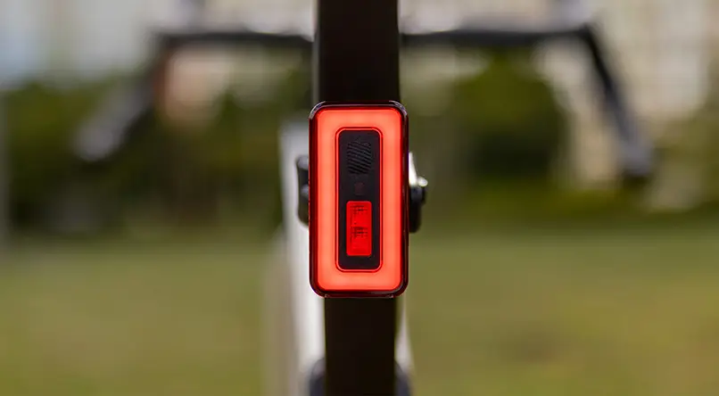 Primer plano de una luz trasera de bicicleta Sigma Nyte 70 LED roja rectangular sujeta a una tija de sillín, con un fondo verde borroso al aire libre.