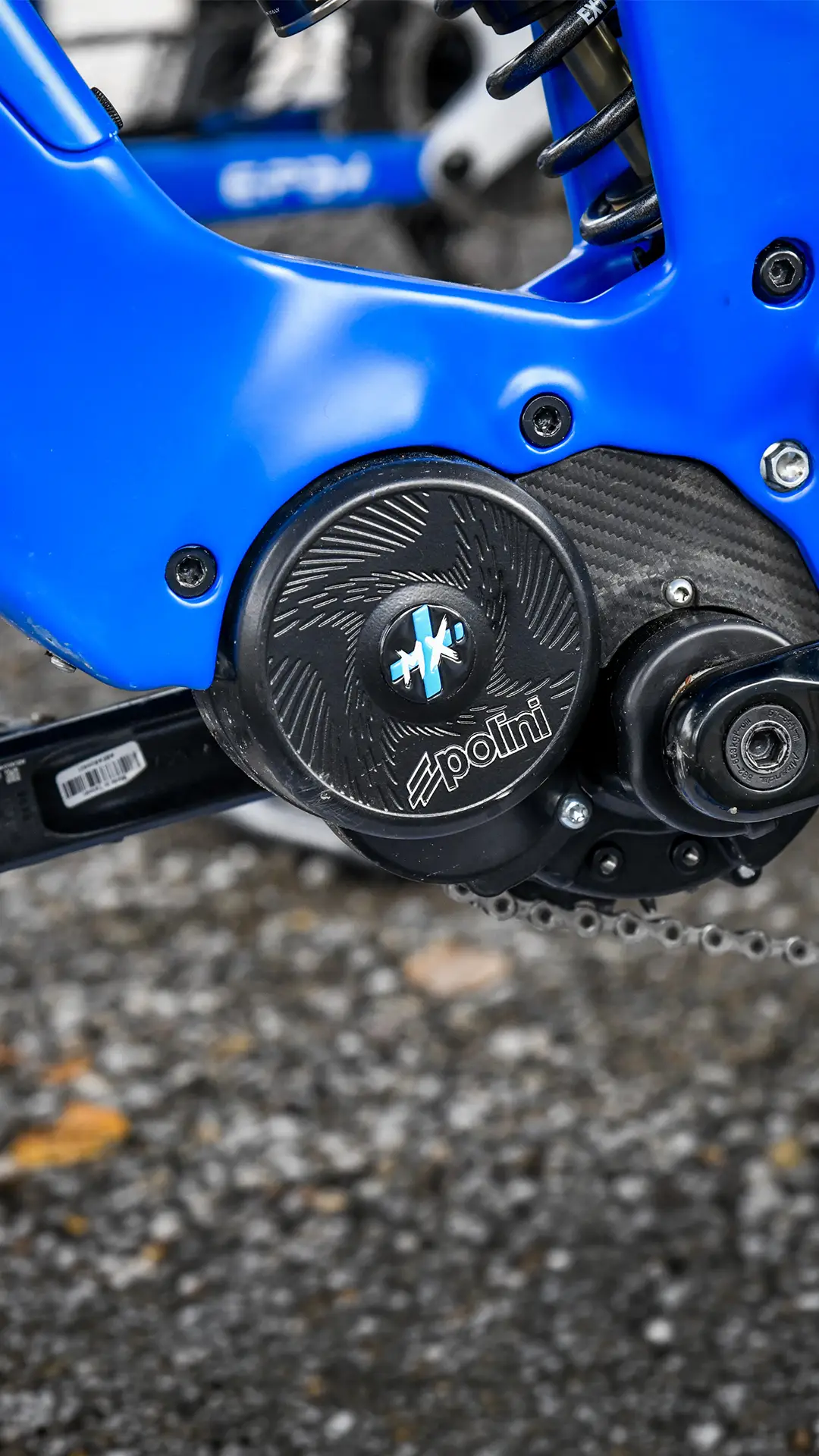 Primer plano de un cuadro de bicicleta azul que muestra un motor Polini negro con pernos visibles y parte de la transmisión por cadena, sobre una superficie de grava.