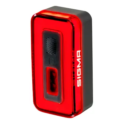 Luz trasera de bicicleta Sigma Nyte 70 rectangular roja y negra con cubierta translúcida y botón central, mostrada en ángulo sobre fondo blanco.