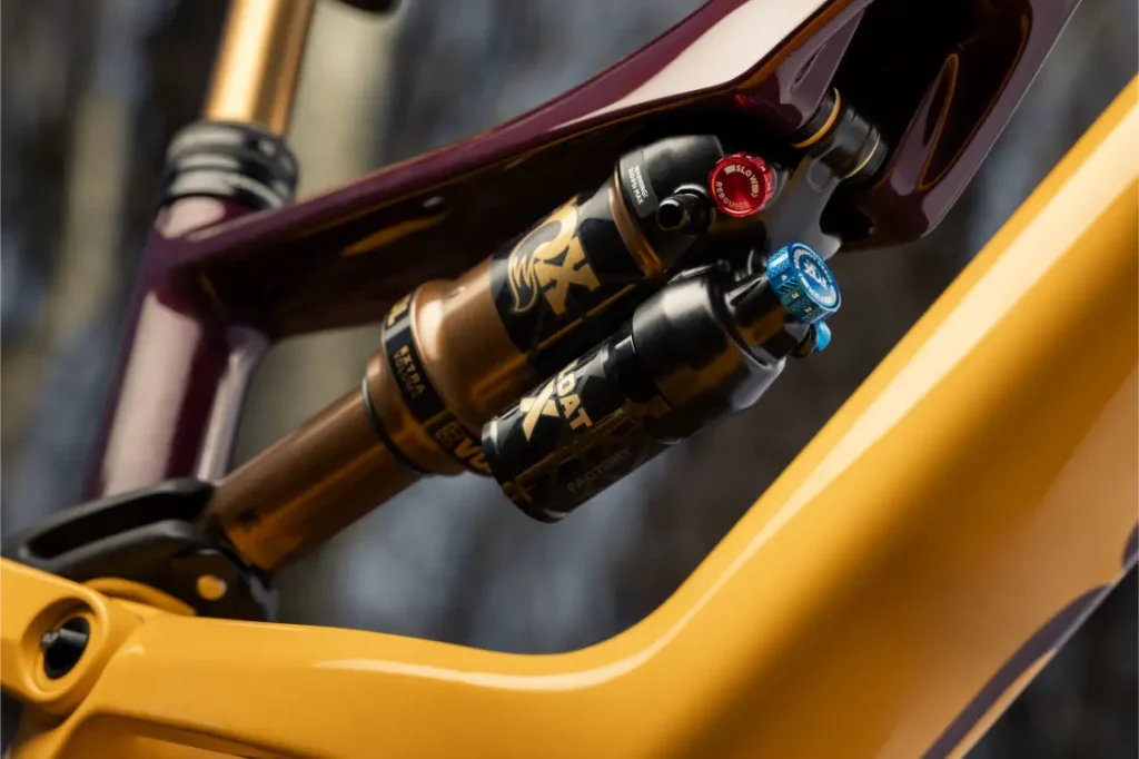 Primer plano del sistema de suspensión trasera de una bicicleta de montaña, con amortiguadores FOX con pomos de ajuste y parte del cuadro de la bicicleta X Factor en amarillo y granate.