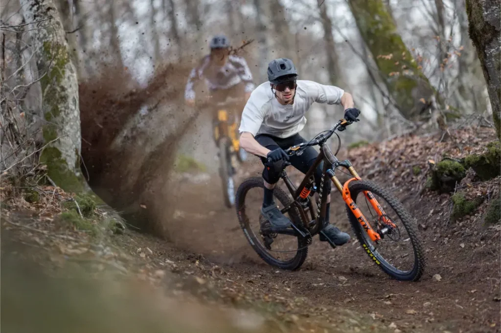 Dos ciclistas de montaña bajan rápidamente por un sendero de tierra del bosque, sus amortiguadores FOX manejando cada curva cerrada mientras la tierra vuela detrás del ciclista que va en cabeza.