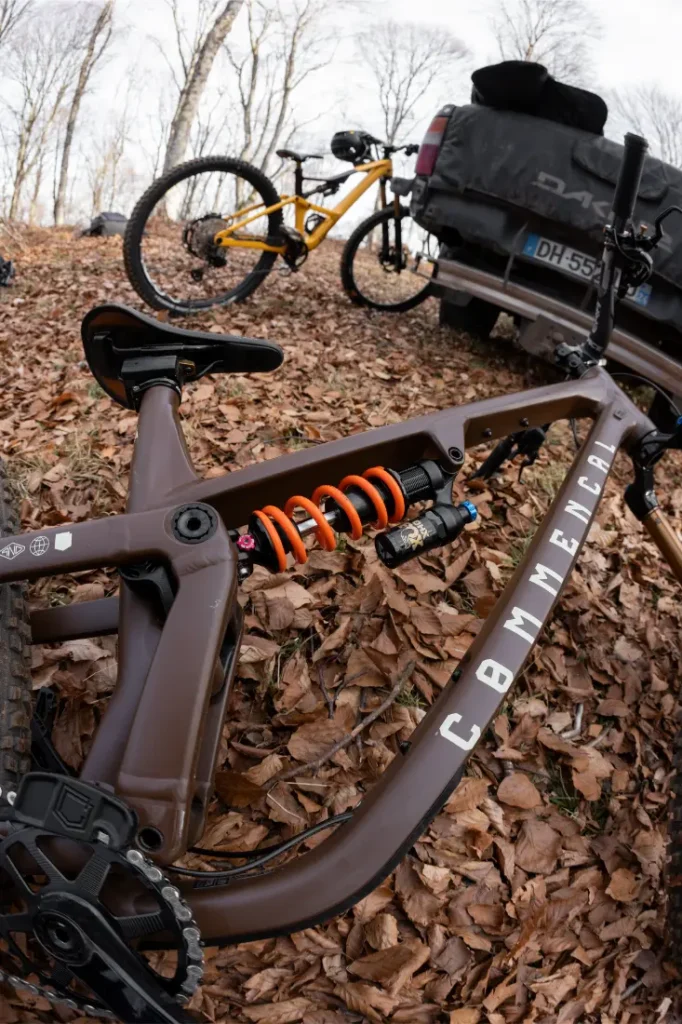 Primer plano de una bicicleta de montaña Commencal marrón con amortiguadores FOX naranjas aparcada sobre hojas secas, otra bicicleta y una camioneta al fondo.