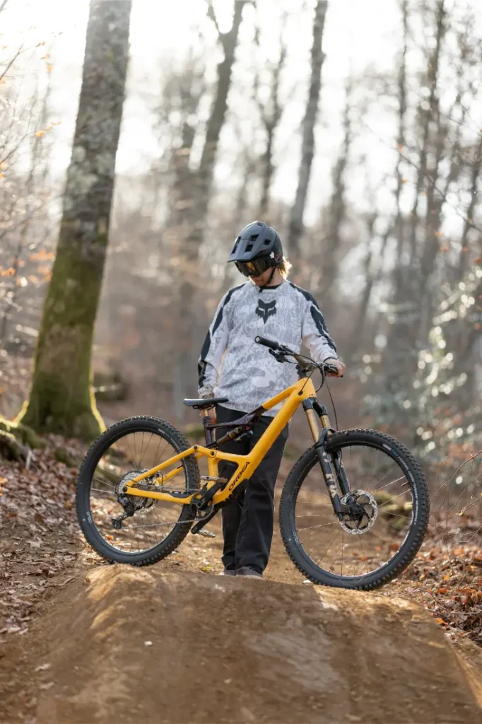 Una persona con casco y ropa de ciclista se encuentra en un sendero de tierra en el bosque, sujetando una bicicleta de montaña amarilla equipada con amortiguadores de primera calidad.