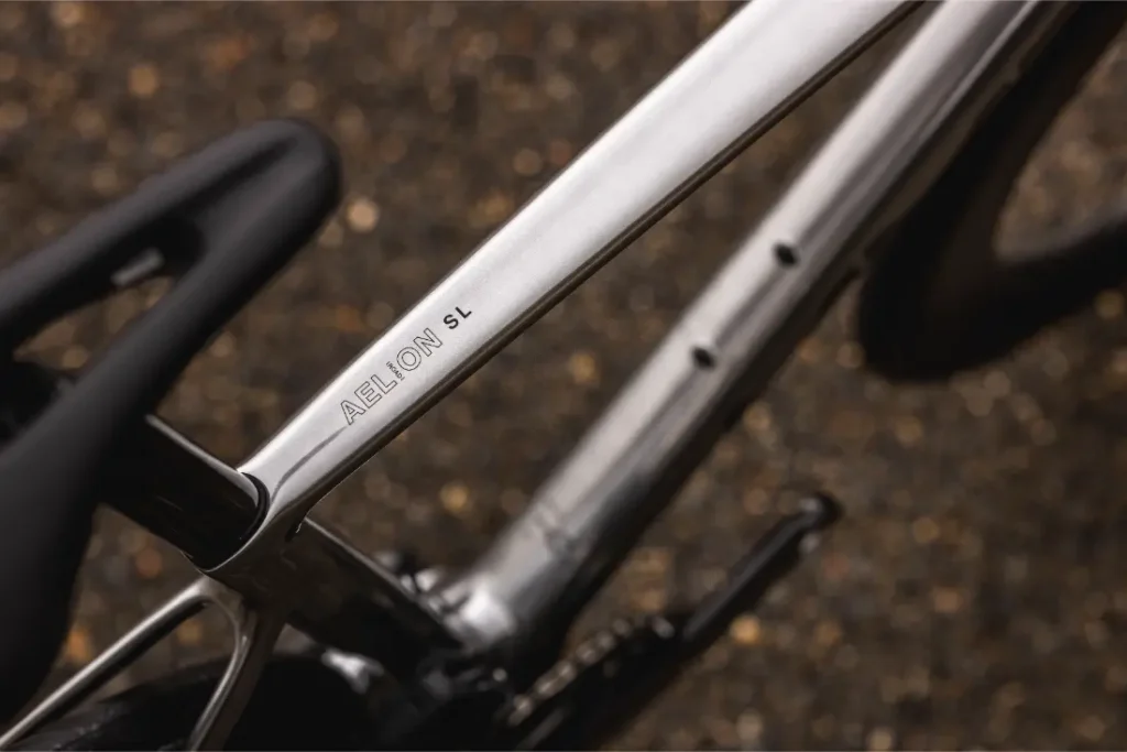 Primer plano de un cuadro de bicicleta metálico etiquetado "AELION SL", con parte del asiento y la rueda trasera visibles sobre un fondo borroso.