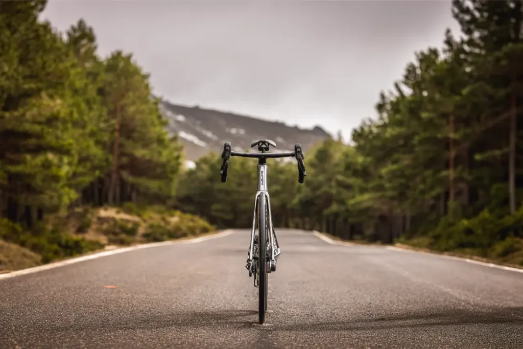 Una bicicleta de carretera MMR Aelion SL se encuentra en posición vertical en el centro de una carretera vacía y asfaltada, rodeada de árboles y montañas bajo un cielo nublado.