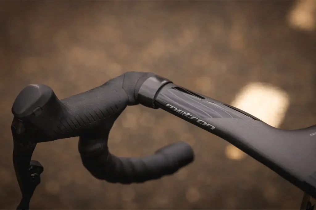 Primer plano del manillar y la potencia integrados de una bicicleta de carretera MMR Aelion negra, con la marca "medron", sobre un fondo marrón difuminado.