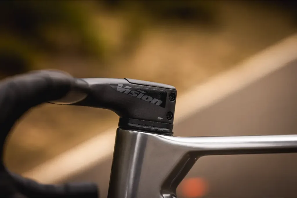 Una mano enguantada agarra la potencia de un manillar Vision en una MMR Aelion SL, sujeta a un cuadro de bicicleta metálico, con una carretera borrosa de fondo: perfecto para cualquier entusiasta de la bici total.