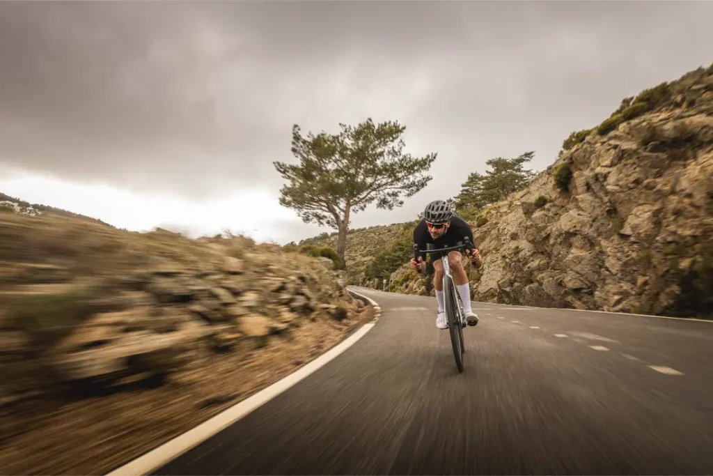Un ciclista con casco conduce una bicicleta de carretera MMR Aelion SL cuesta abajo por una sinuosa carretera de montaña, rodeado de un paisaje rocoso y un cielo nublado.