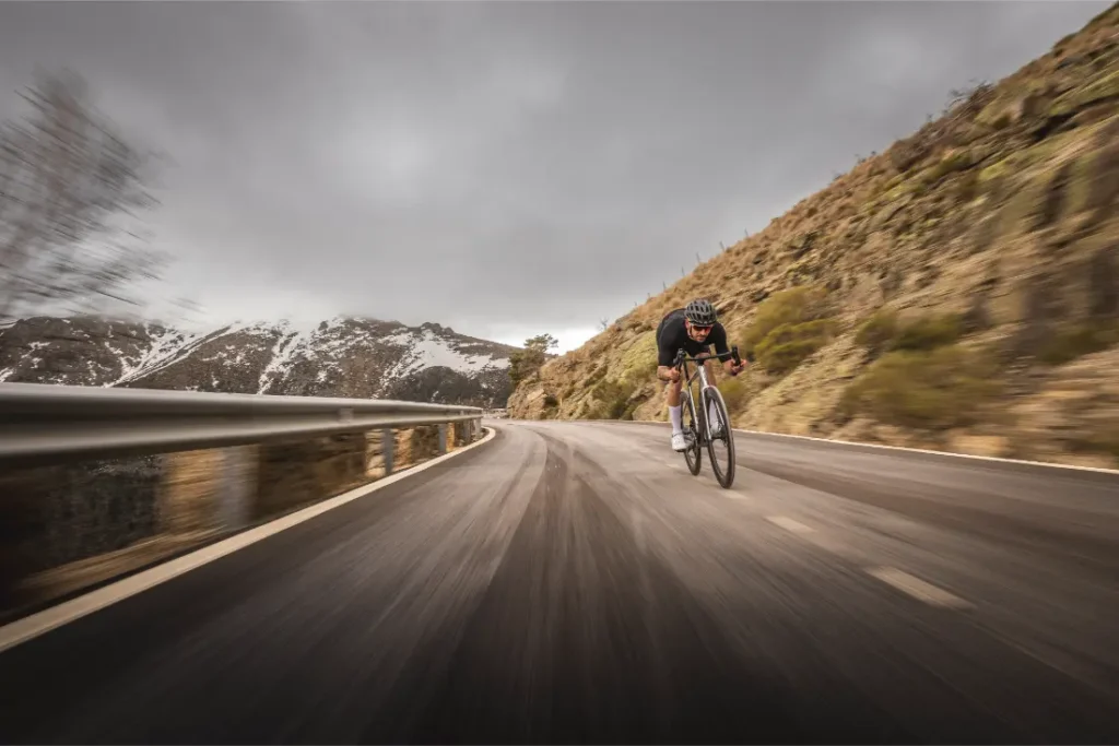 Un ciclista desciende por una curva carretera de montaña bajo un cielo nublado en una Aelion SL, con los picos nevados visibles al fondo.