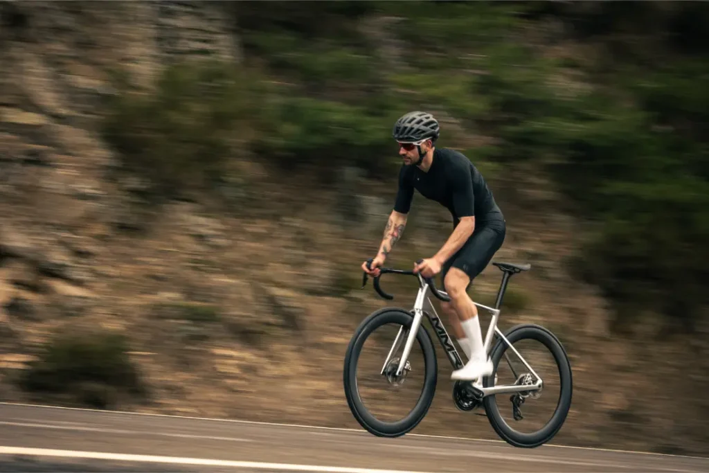 Una persona con casco y ropa de ciclismo monta en una bicicleta de carretera MMR Aelion SL por una carretera asfaltada con un fondo natural difuminado, mostrando las prestaciones del equipamiento bici total.