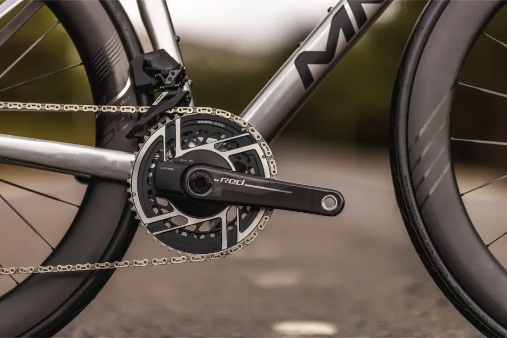 Primer plano de unas bielas y un plato de bicicleta Aelion SL fijados a un cuadro de bicicleta plateado, con parte de la rueda trasera y el neumático visibles.