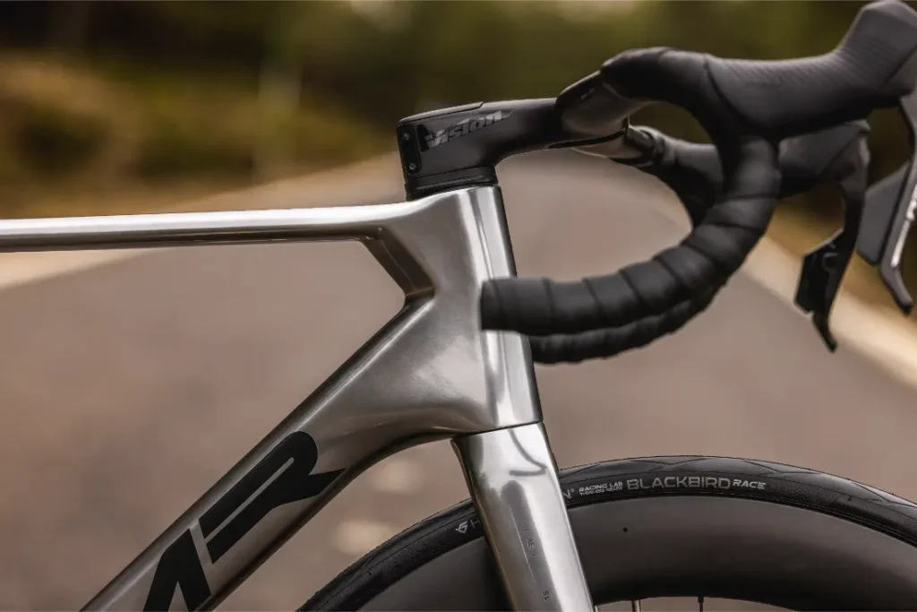 Primer plano del manillar, el cuadro y la rueda delantera de una bicicleta MMR Aelion en una carretera asfaltada vacía con un fondo natural asturiano difuminado.
