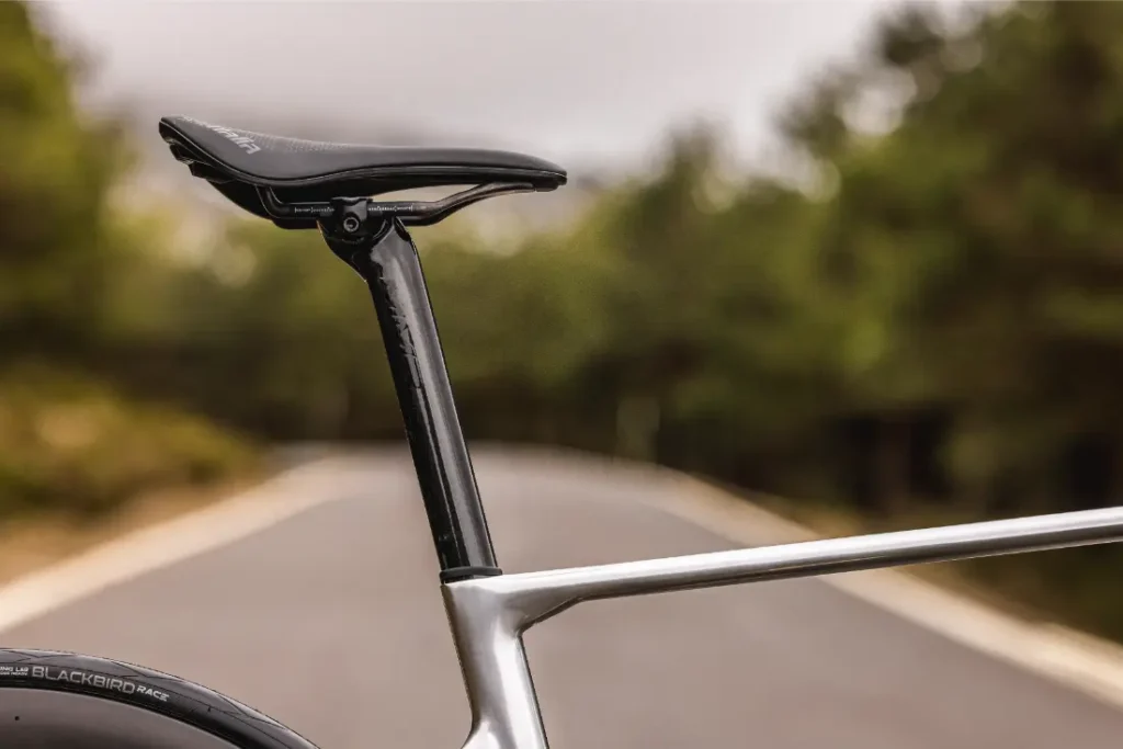 Primer plano del sillín de una bicicleta MMR Aelion, la tija del sillín y parte del cuadro con una carretera exterior difuminada y árboles al fondo-un toque de bici total con acento asturiano.