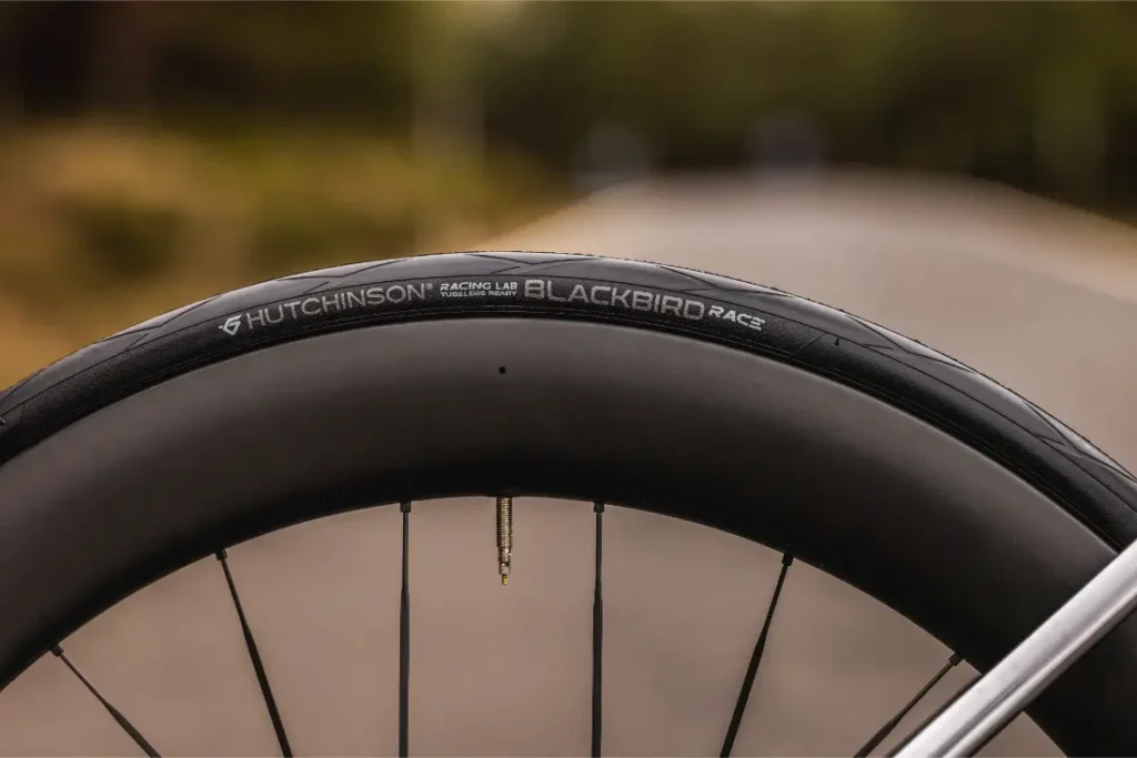 Primer plano de una rueda de bicicleta MMR Aelion con un neumático Hutchinson Racing Lab Blackbird, con una carretera borrosa y vegetación de fondo.