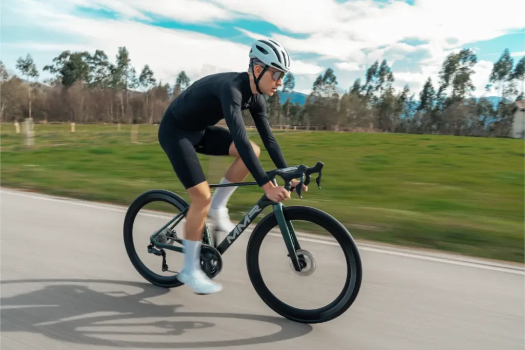 Un ciclista con atuendo negro y casco blanco conduce una bicicleta de carretera MMR Aelion SL por una carretera asfaltada con hierba verde y árboles de fondo, mostrando el espíritu de la bici asturiana.