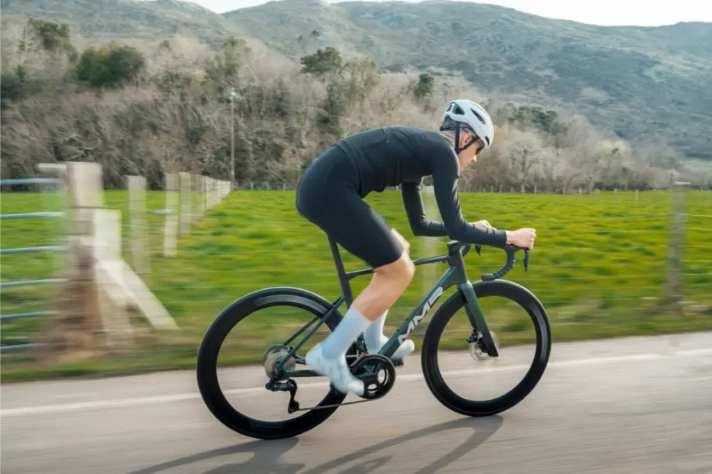 Un ciclista con casco y ropa de ciclismo conduce una bicicleta de carretera MMR Aelion SL por una carretera rural con verdes campos y colinas de fondo, encarnando el verdadero espíritu de la bici total.
