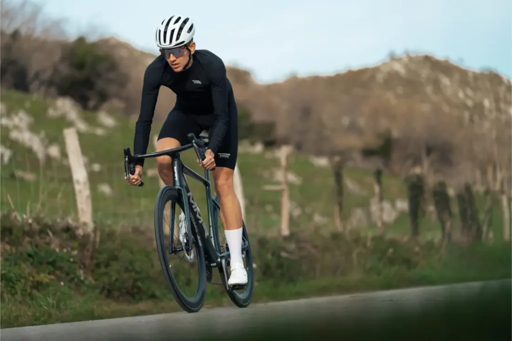 Un ciclista con atuendo negro, casco blanco y gafas de sol conduce una bicicleta de carretera MMR Aelion SL por un camino rural con colinas y árboles de fondo.