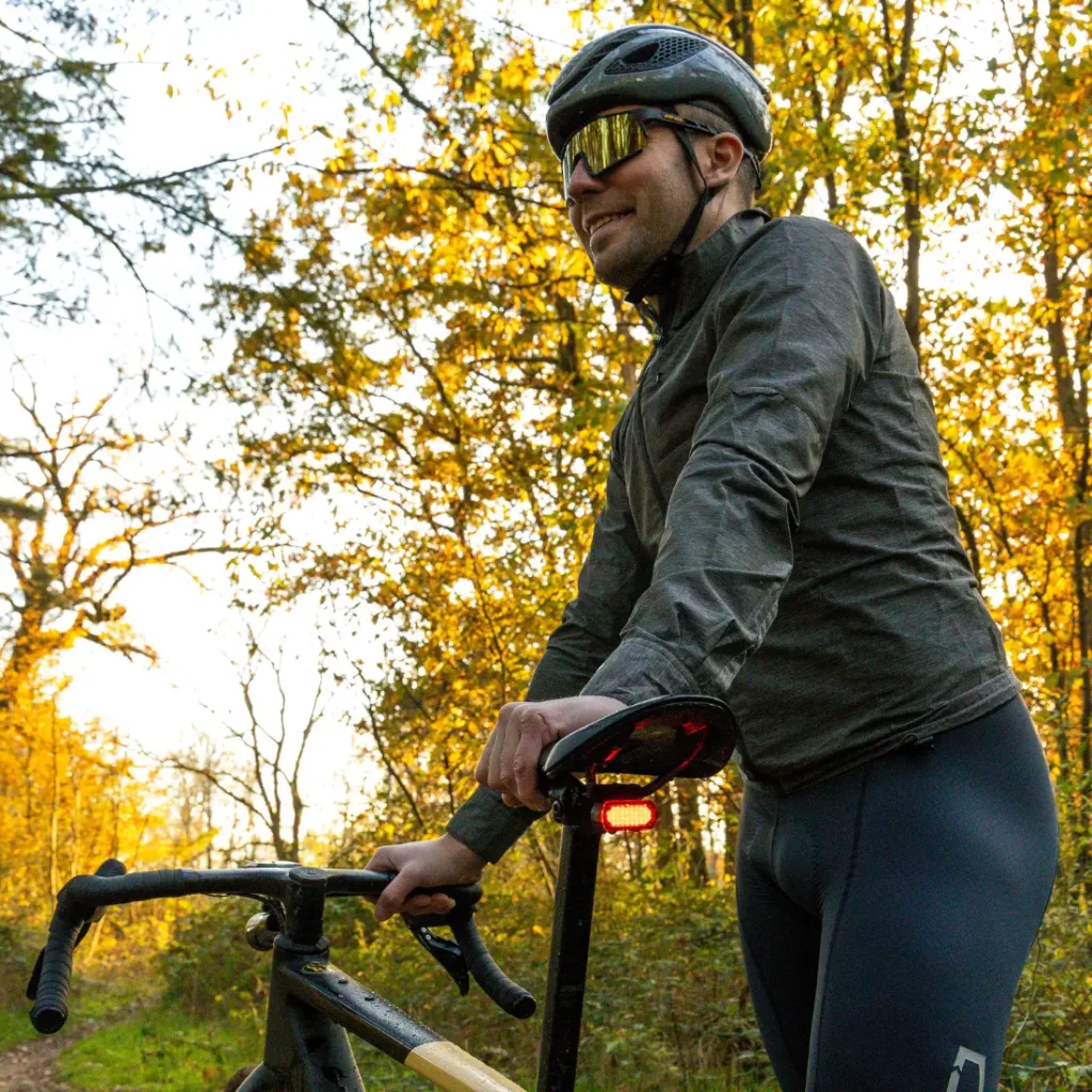 Un ciclista con casco, gafas de sol y ropa oscura con el CLIP SIGMA NYTE colocado se sitúa junto a una bicicleta en un camino forestal bordeado de follaje otoñal.