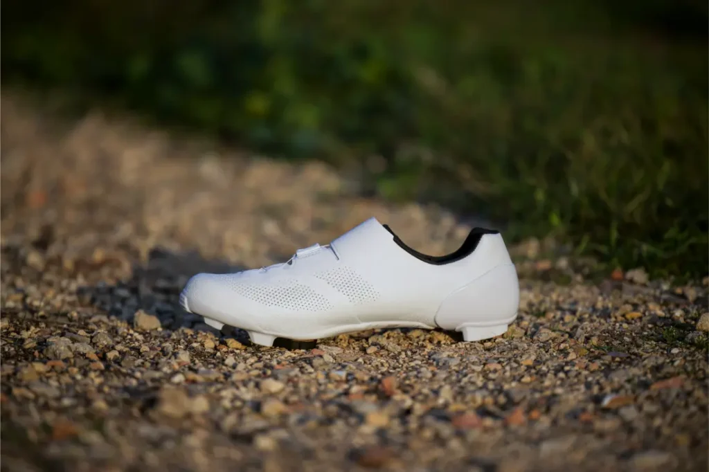 Unas Zapatillas Shimano RX910 S-Phyre blancas de ciclismo descansan sobre un camino de grava con vegetación en el fondo borroso.