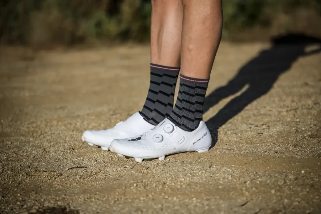 Una persona está de pie en un camino de grava, con calcetines a rayas negras y grises y zapatillas de ciclismo RX910 S-Phyre Zapatillas Shimano blancas.