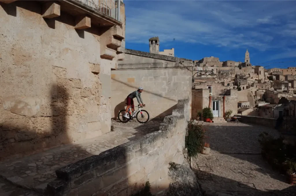 Un ciclista que lleva ASSOS MILLE GT S11 disfruta del confort inteligente mientras recorre un estrecho sendero de piedra en una antigua ciudad en lo alto de una colina con edificios de color canela bajo un cielo azul.