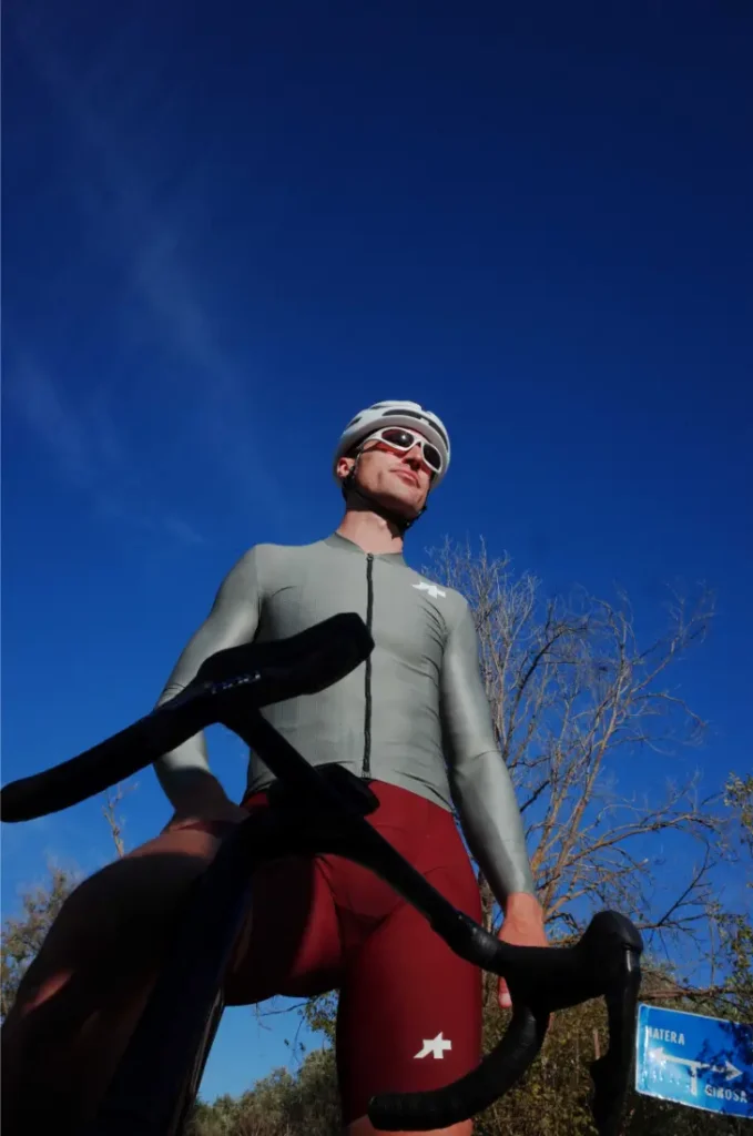 Un ciclista con un maillot gris ASSOS MILLE GT S11 y un culotte rojo junto a una bicicleta bajo un cielo azul claro, encarnando la evolución real y el confort inteligente con árboles de fondo.