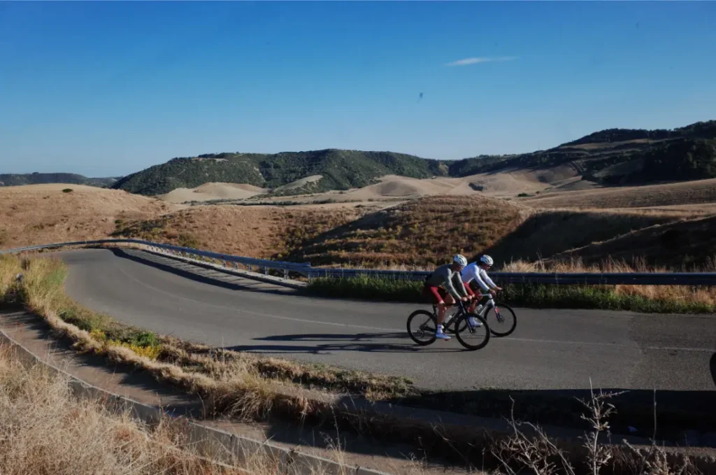 Dos ciclistas recorren una carretera asfaltada a través de un paisaje montañoso y seco bajo un cielo azul despejado, mostrando la evolución real y el confort inteligente de la equipación ASSOS MILLE GT S11.