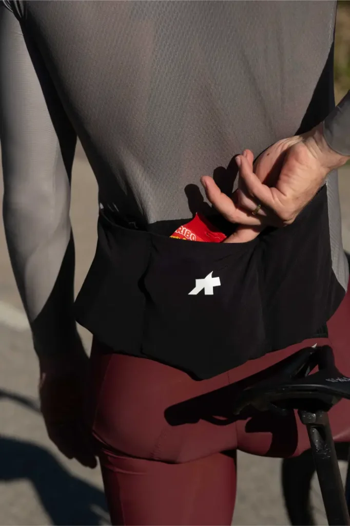 Un ciclista con un maillot ASSOS MILLE GT S11 mete la mano en el bolsillo trasero y saca un gel energético junto a una bicicleta, mostrando la evolución real y el confort inteligente en cada salida.
