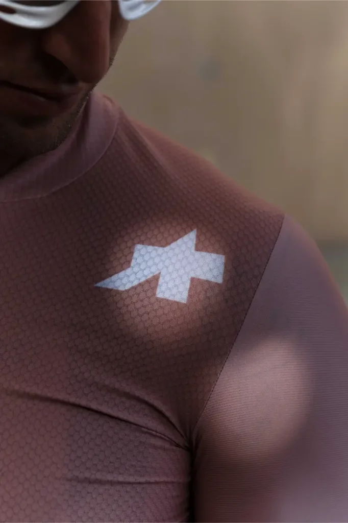 Primer plano de una persona que lleva un maillot de ciclismo ASSOS MILLE GT S11 de color malva con un logotipo geométrico blanco en el hombro, que muestra la evolución real y el confort inteligente; sólo se ven parte de la cara y la parte superior del torso.