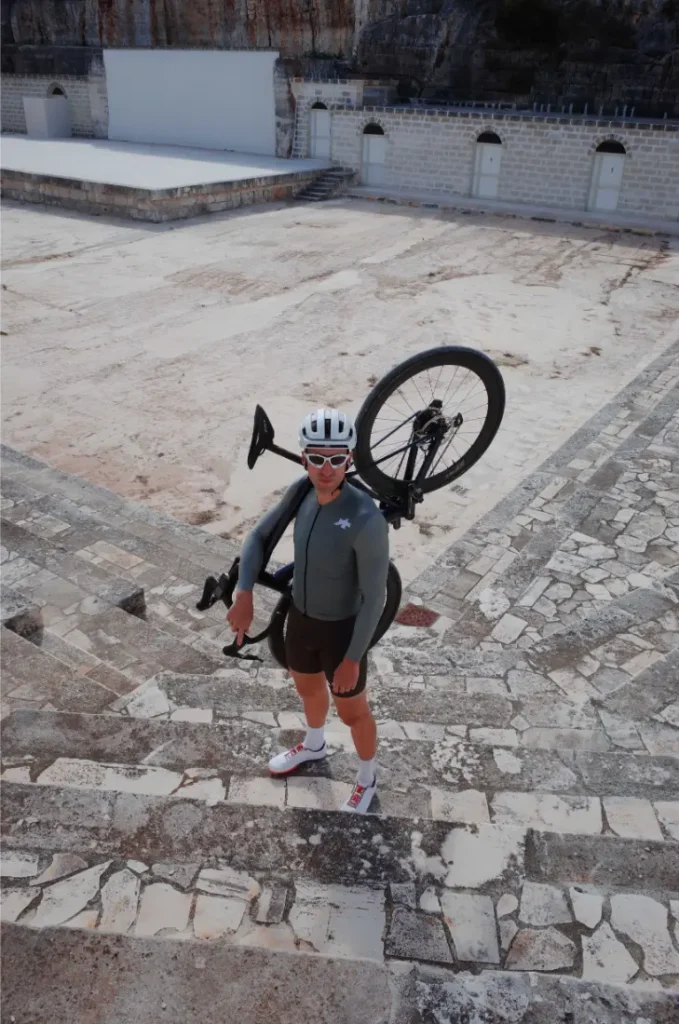 Un ciclista ataviado con una equipación ASSOS MILLE GT S11, casco y gafas de sol, de pie sobre unos escalones de piedra y con una bicicleta al hombro, en un escenario de piedra vacío y al aire libre, muestra a la perfección la evolución real y el confort inteligente.