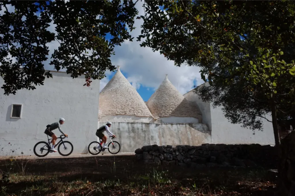 Dos ciclistas con equipamiento ASSOS MILLE GT S11 pasan por delante de tradicionales casas trulli blancas con tejados cónicos, enmarcadas por árboles y un muro de piedra bajo un cielo parcialmente nublado, disfrutando de confort inteligente en su viaje.