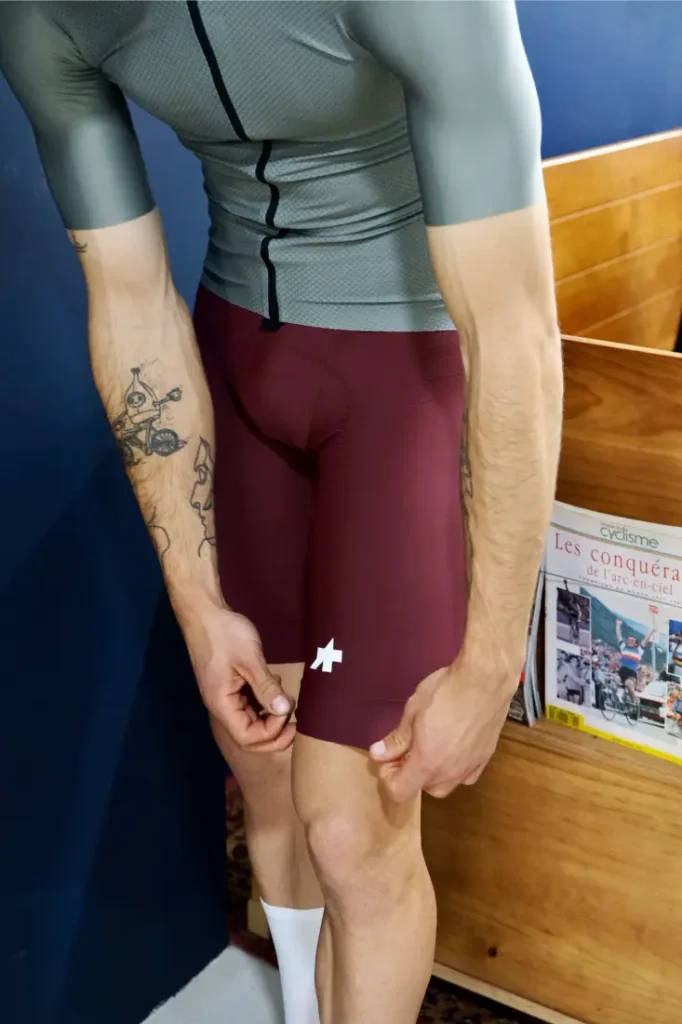 Una persona vestida de ciclista se ajusta el culotte granate ASSOS MILLE GT S11 junto a un estante de madera con revistas de ciclismo, mostrando confort inteligente y evolución real en sus elecciones de equipamiento.