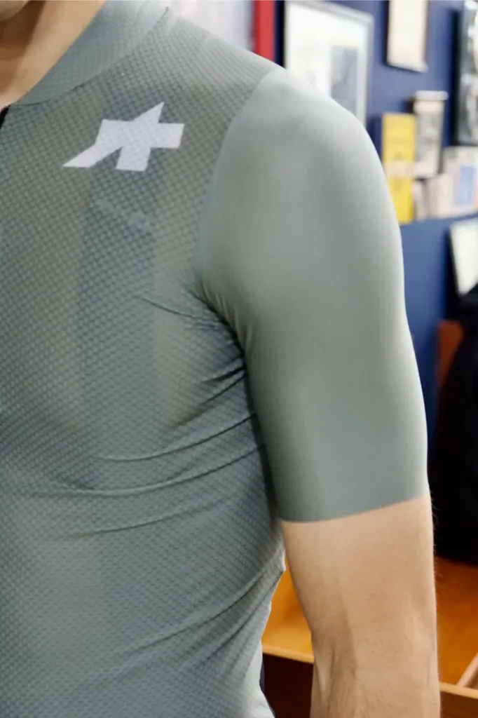 Persona que lleva una camiseta deportiva ASSOS MILLE GT S11 gris, ajustada y de manga corta, con un estampado texturizado y un logotipo blanco en el hombro izquierdo; el fondo incluye imágenes enmarcadas difuminadas. Perfecto para quienes buscan confort inteligente.